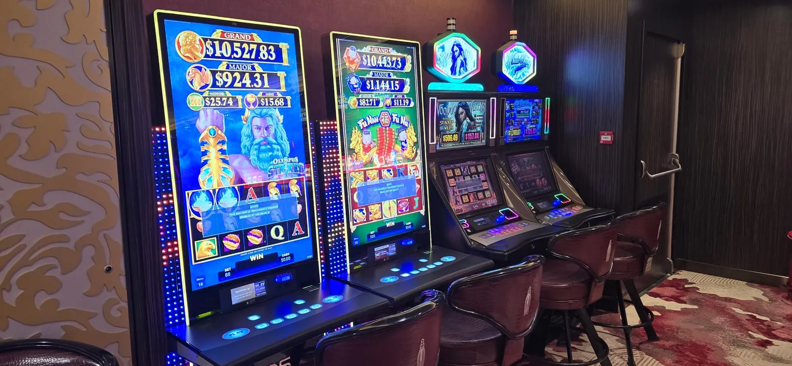 Two tall portrait-screen slot machines, Olympus Strikes with a Zeus figure and Fu Nan Fu Nu with Chinese artwork, standing beside two shorter cabinets topped with illuminated Voodoo Vixens and winter-themed signs.