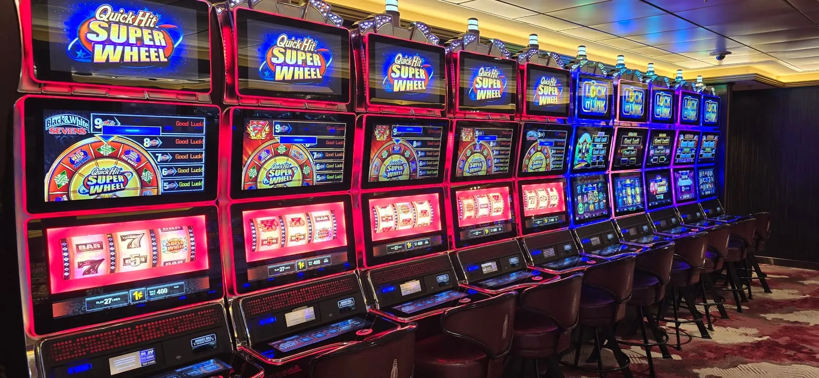 A long row of Quick Hit Super Wheel cabinets lit in red, giving way to a bank of Lock It Link machines lit in blue further down the row.