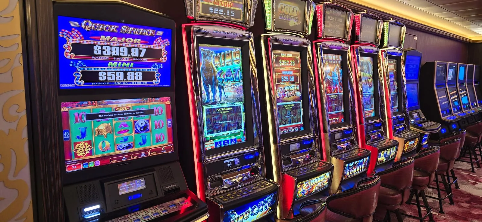 A Konami Quick Strike machine in the foreground showing Major and Mini jackpots with their posted ranges, followed by a receding row of Ainsworth cabinets including King Cat, Dragon Lines and Rumble Rumble.